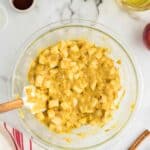 A glass bowl with chopped apples mixed into yellow batter, surrounded by ingredients including oil, vanilla, cinnamon sticks, and powdered sugar on a marble countertop.
