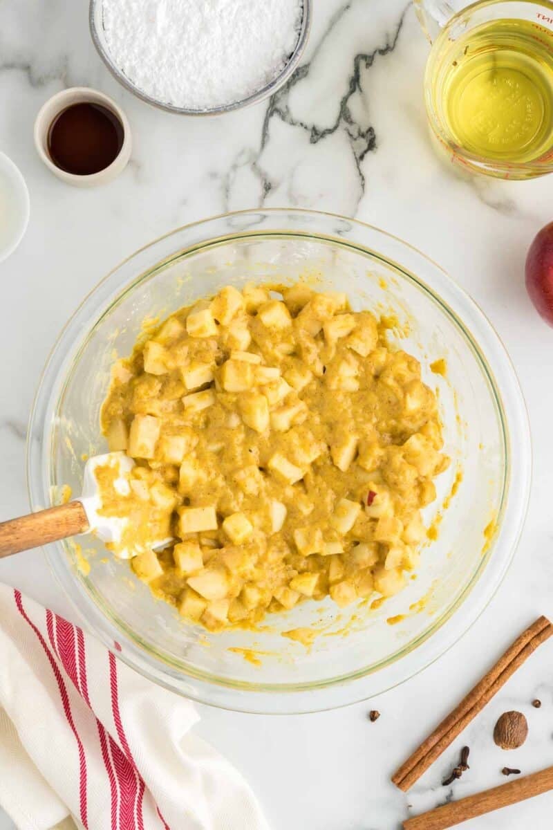 A glass bowl with chopped apples mixed into yellow batter, surrounded by ingredients including oil, vanilla, cinnamon sticks, and powdered sugar on a marble countertop.