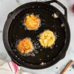 Three apple fritters frying in hot oil in a black skillet on a marble countertop, with a red-striped towel, cinnamon sticks, apples, and small bowls nearby.