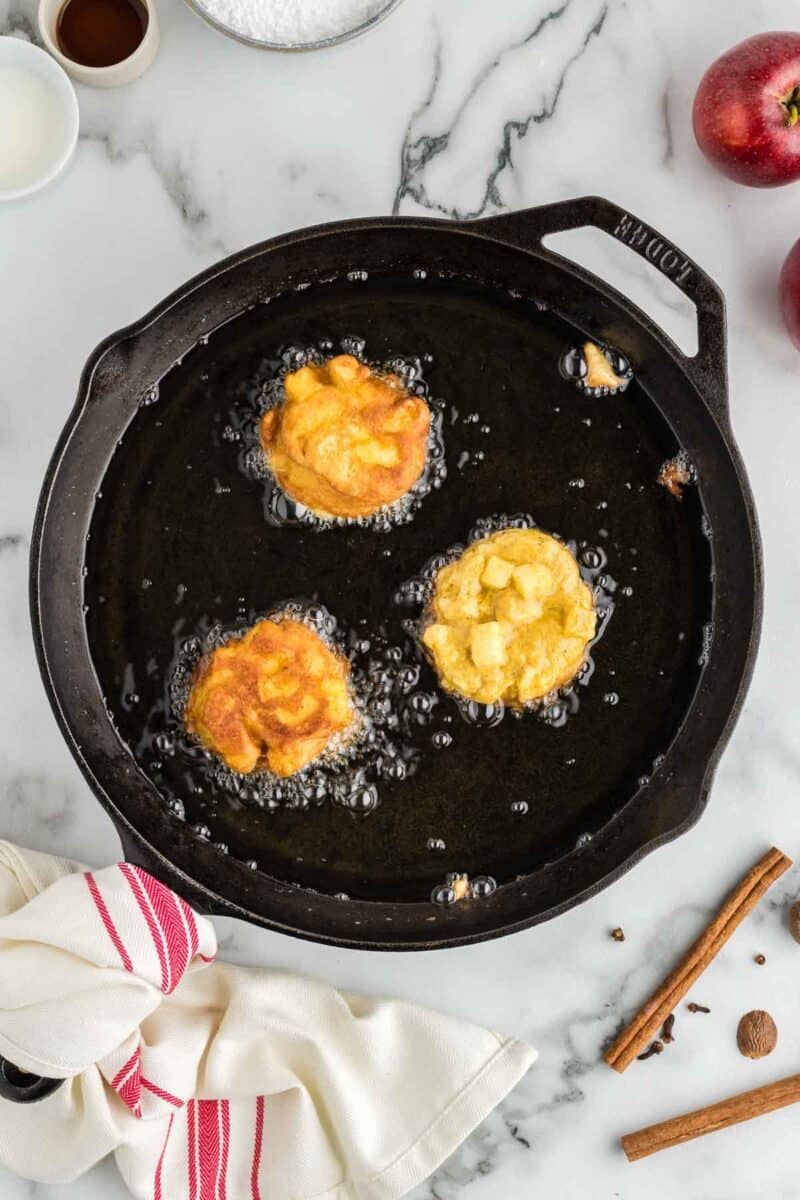 Three apple fritters frying in hot oil in a black skillet on a marble countertop, with a red-striped towel, cinnamon sticks, apples, and small bowls nearby.