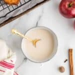 A bowl of light-colored glaze with a gold spoon sits on a marble surface near a red apple, cinnamon stick, nutmeg, cloves, a towel, and baked goods on a cooling rack.