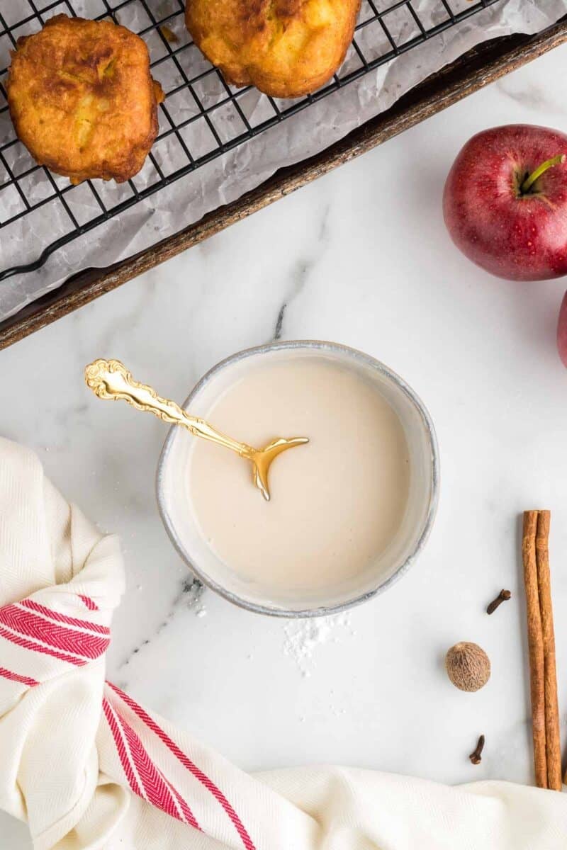 A bowl of light-colored glaze with a gold spoon sits on a marble surface near a red apple, cinnamon stick, nutmeg, cloves, a towel, and baked goods on a cooling rack.