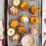 A baking tray with nine apple fritters, some topped with white glaze, sits on a wire rack. Sliced apples and a bowl of glaze with a spoon are also on the tray.