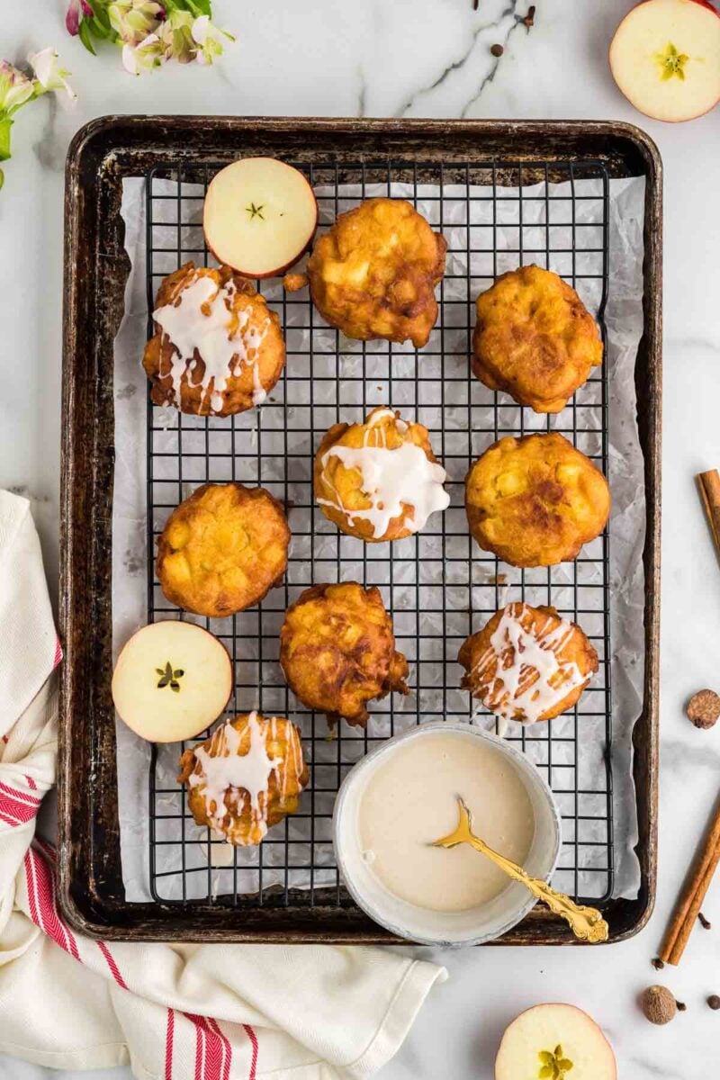 A baking tray with nine apple fritters, some topped with white glaze, sits on a wire rack. Sliced apples and a bowl of glaze with a spoon are also on the tray.