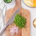 Chopped jalapeños and red onions on a wooden cutting board with a knife, surrounded by bowls of cilantro, shredded cheese, chips, and a white-striped towel.