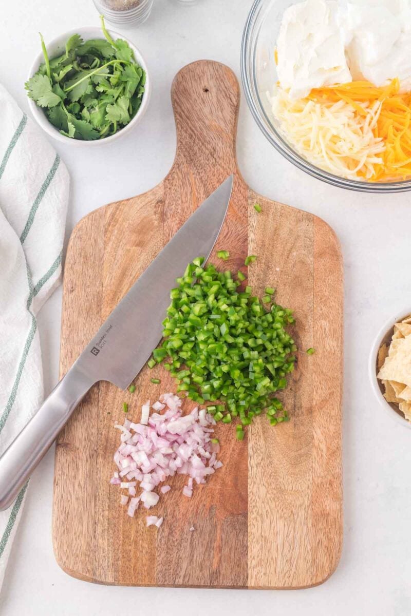 Chopped jalapeños and red onions on a wooden cutting board with a knife, surrounded by bowls of cilantro, shredded cheese, chips, and a white-striped towel.