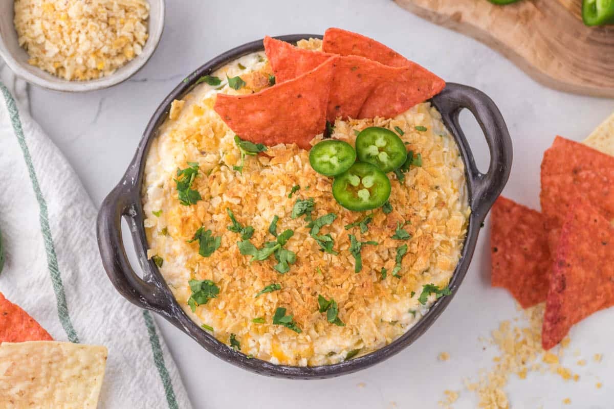 A bowl of creamy dip topped with crushed chips, chopped herbs, sliced jalapeños, and red tortilla chips, placed on a white surface with additional chips nearby.
