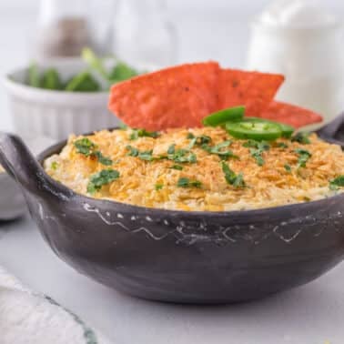 A black bowl filled with a baked cheese dip, topped with chopped herbs, sliced jalapenos, and two red tortilla chips, sits on a light-colored table with snacks in the background.