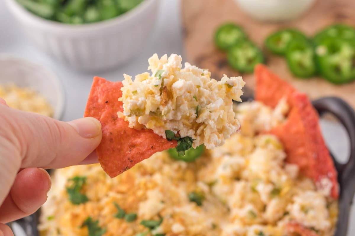 A hand holding a red tortilla chip topped with cheesy, crumbly dip; a bowl of dip and sliced jalapeños are visible in the background.