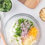 A glass bowl contains sour cream, shredded cheddar and Monterey Jack cheese, chopped jalapeño, and diced red onion. Nearby are cilantro, tortilla chips, and a cutting board with a knife.