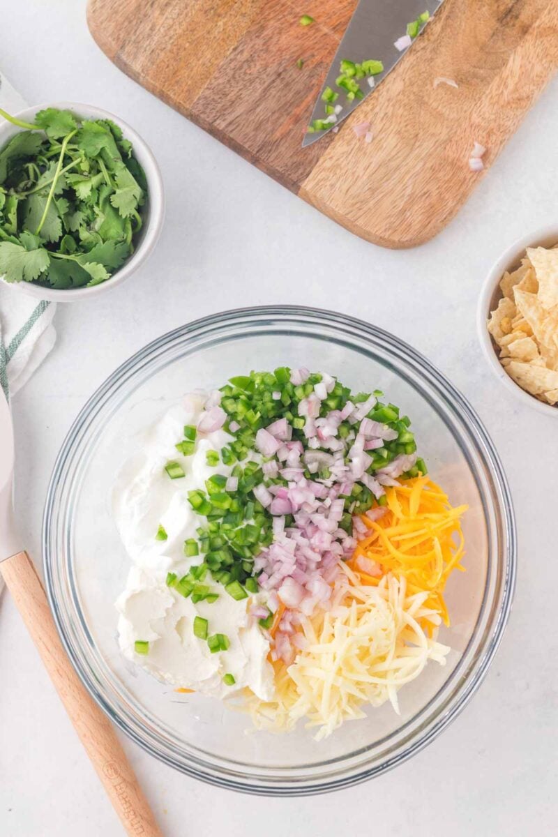 A glass bowl contains sour cream, shredded cheddar and Monterey Jack cheese, chopped jalapeño, and diced red onion. Nearby are cilantro, tortilla chips, and a cutting board with a knife.