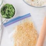 A plastic bag with crushed crackers, a rolling pin, a bowl of fresh cilantro, and a bowl of creamy mixture on a white surface.