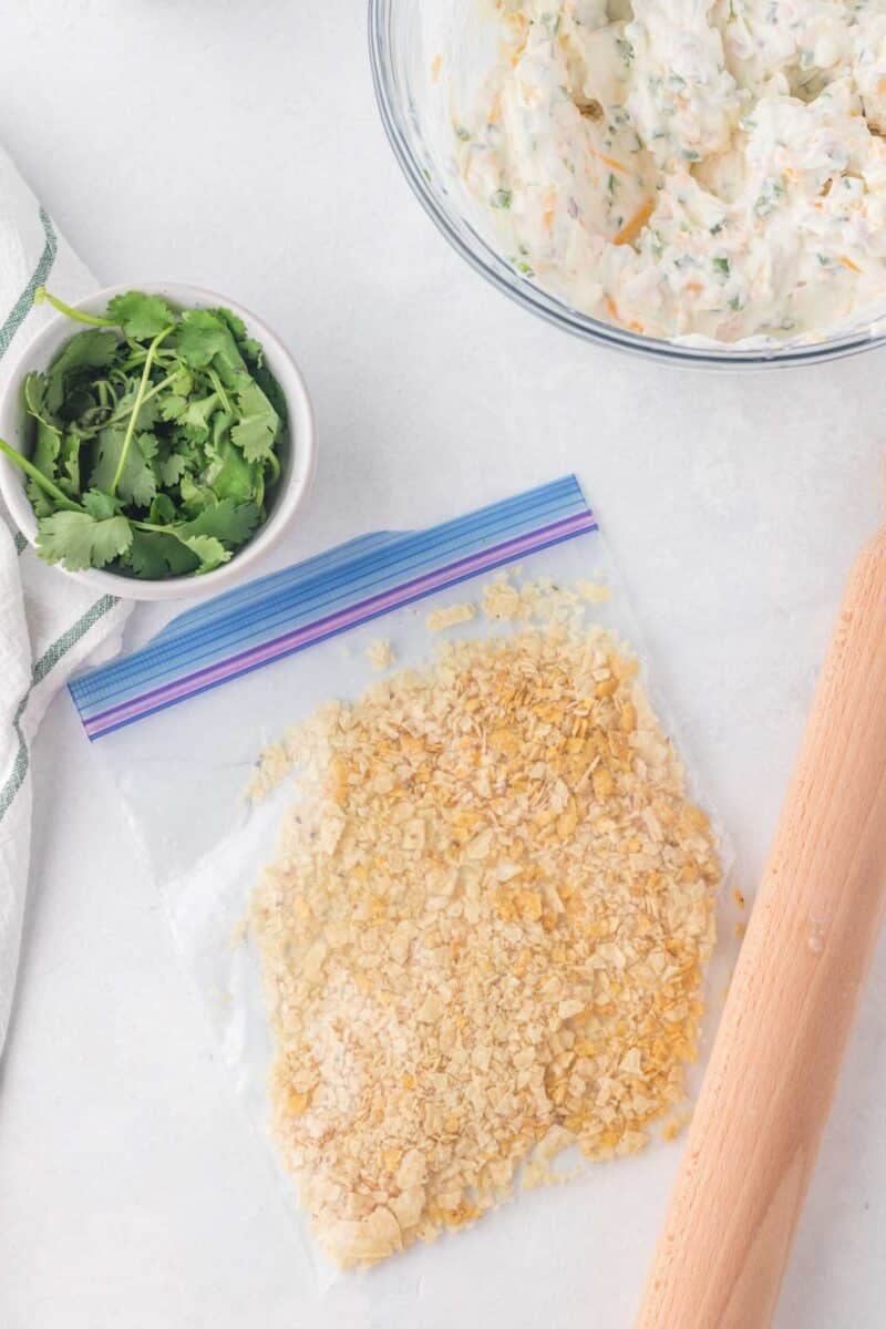 A plastic bag with crushed crackers, a rolling pin, a bowl of fresh cilantro, and a bowl of creamy mixture on a white surface.