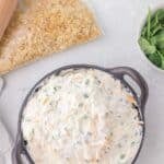 A bowl of creamy dip with chopped vegetables and shredded cheese sits beside a bag of crushed crackers, a rolling pin, and a bowl of fresh cilantro on a white surface.