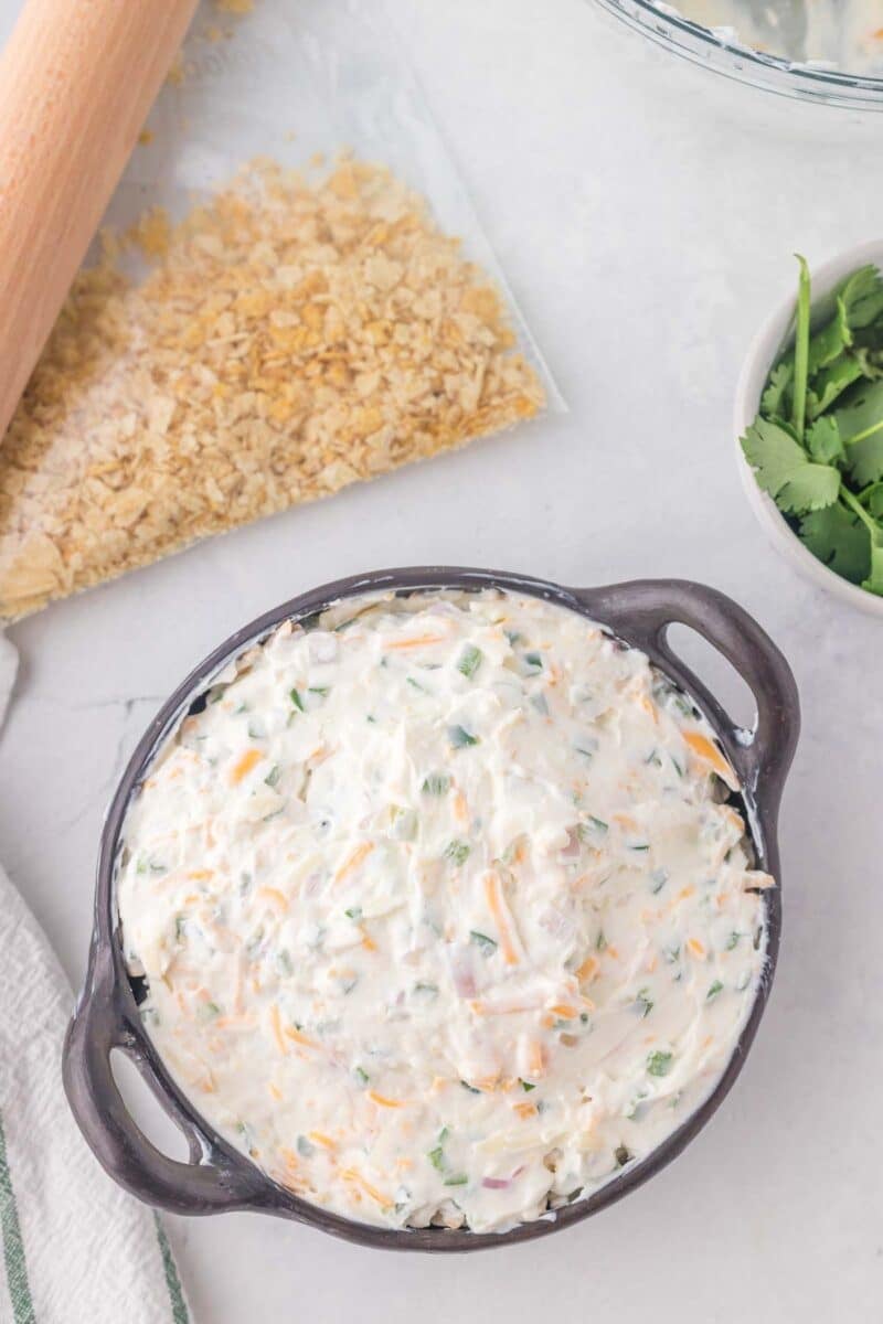 A bowl of creamy dip with chopped vegetables and shredded cheese sits beside a bag of crushed crackers, a rolling pin, and a bowl of fresh cilantro on a white surface.