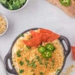 A bowl of cheesy dip topped with crushed chips, cilantro, jalapeño slices, and three triangular chips; cilantro, jalapeño slices, and crumbs are nearby.