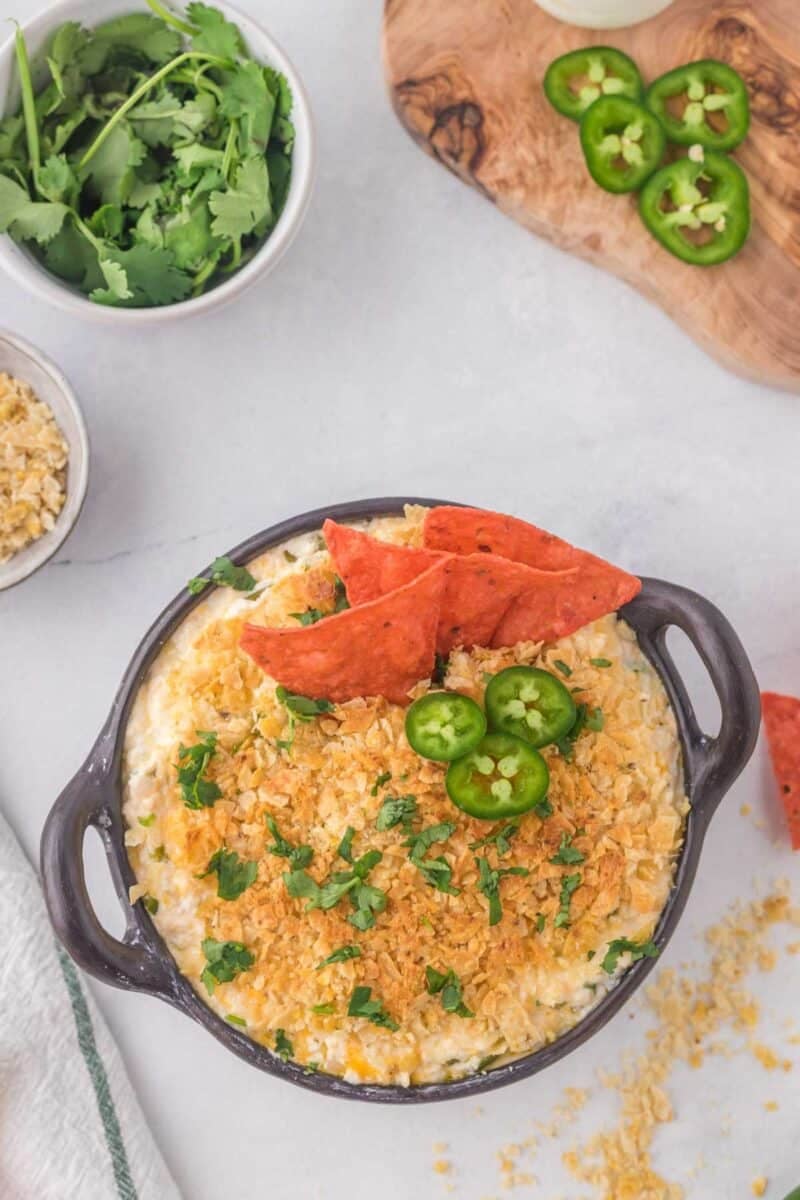 A bowl of cheesy dip topped with crushed chips, cilantro, jalapeño slices, and three triangular chips; cilantro, jalapeño slices, and crumbs are nearby.
