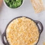 A round baking dish filled with a creamy casserole topped with crushed crackers, next to a small bowl of fresh cilantro and a partially opened bag of crackers.
