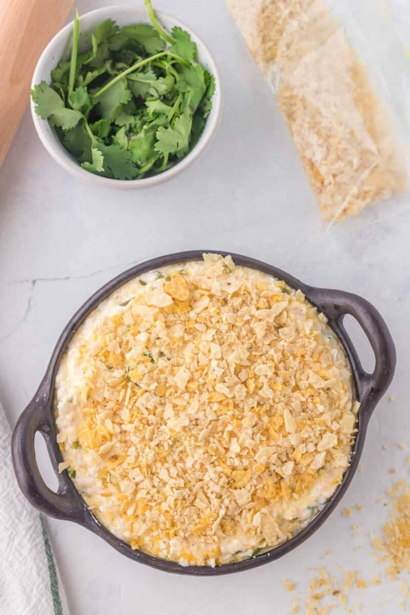 A round baking dish filled with a creamy casserole topped with crushed crackers, next to a small bowl of fresh cilantro and a partially opened bag of crackers.