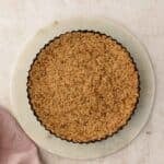A baked crumb crust in a fluted tart pan, resting on a round marble board over a light stone surface with a pink linen napkin nearby.
