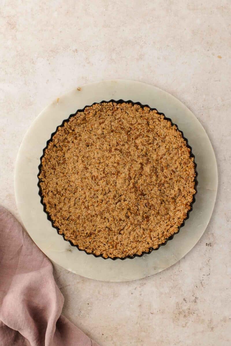 A baked crumb crust in a fluted tart pan, resting on a round marble board over a light stone surface with a pink linen napkin nearby.
