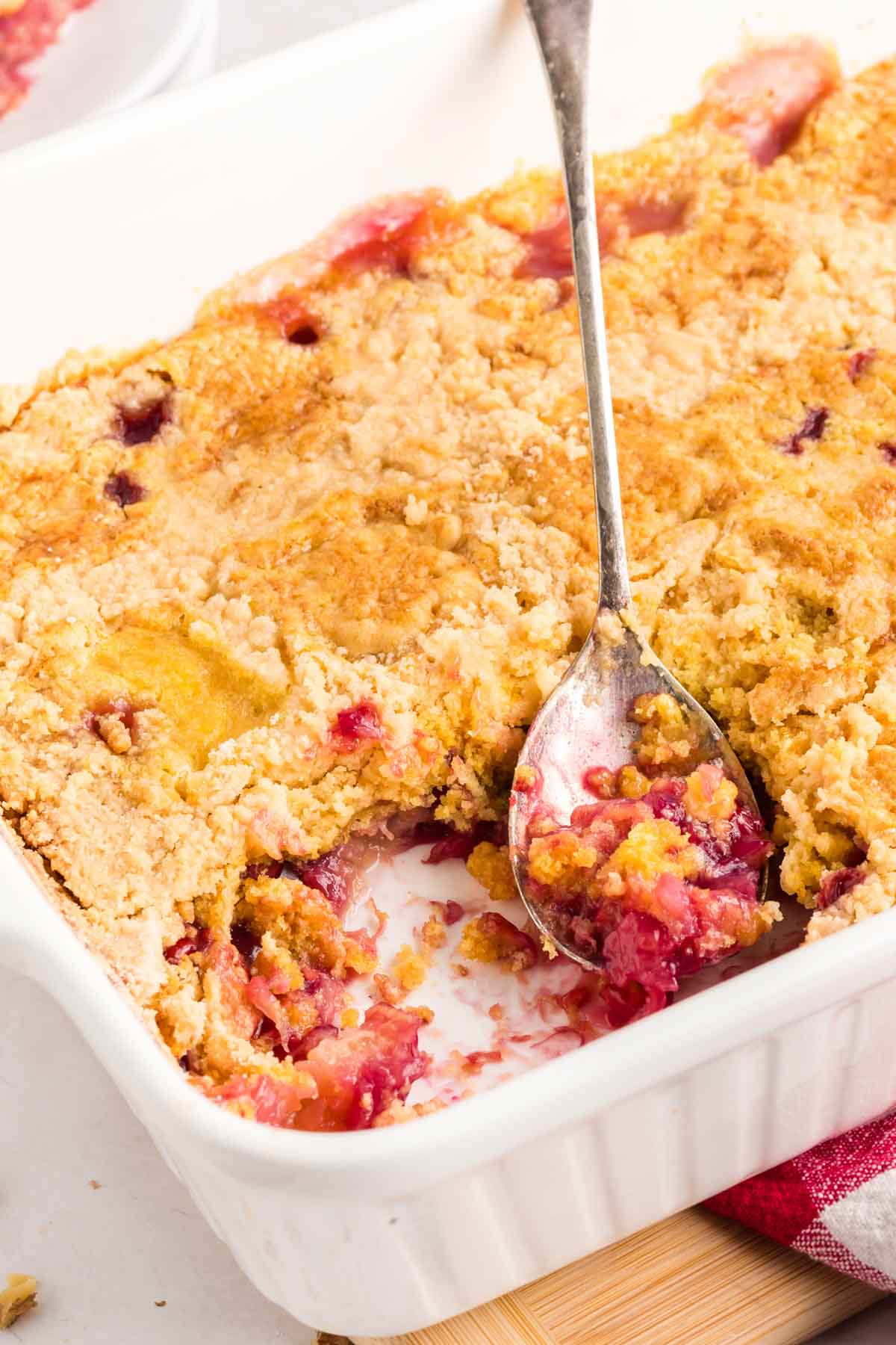 A close-up of a partially served cherry pineapple dump cake in a white baking dish with a spoon scooping out a portion.