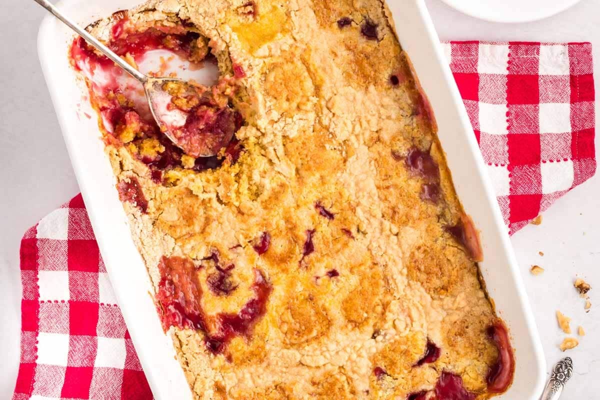 A rectangular baking dish with a partially served cherry pineapple dump cake, a spoon resting in one corner, on top of a red and white checkered cloth.