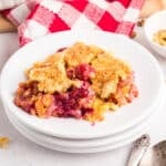 A serving of cherry pineapple dump cake with a golden topping on a white plate, accompanied by a spoon, a small bowl of chopped nuts, and a red and white checkered cloth.