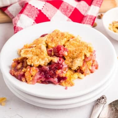 A serving of cherry pineapple dump cake with a golden topping on a white plate, accompanied by a spoon, a small bowl of chopped nuts, and a red and white checkered cloth.