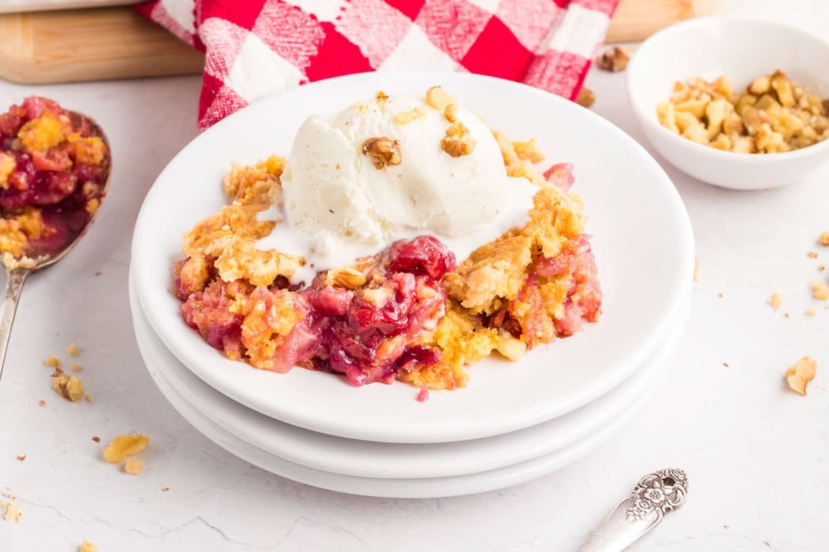 A serving of cherry pineapple dump cake topped with vanilla ice cream and chopped walnuts on a white plate, with a red and white napkin in the background.