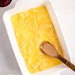 Rectangular baking dish with crushed pineapple and melted butter being mixed with a wooden spoon, surrounded by small bowls of chopped nuts and cherries on a white surface.