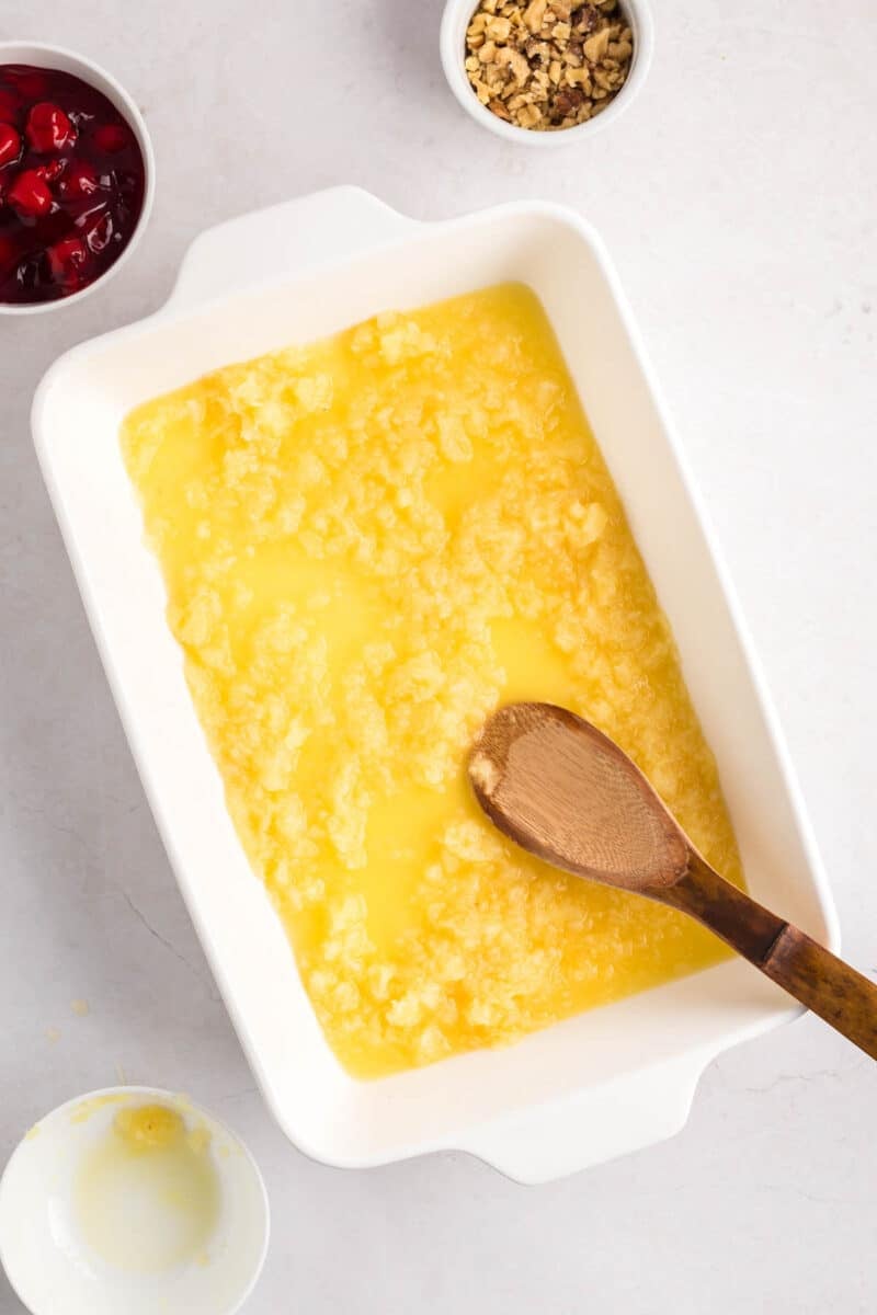 Rectangular baking dish with crushed pineapple and melted butter being mixed with a wooden spoon, surrounded by small bowls of chopped nuts and cherries on a white surface.
