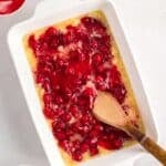 A white baking dish with cherry pie filling and pineapple mixture being stirred with a wooden spoon, surrounded by bowls of chopped nuts and whipped topping.