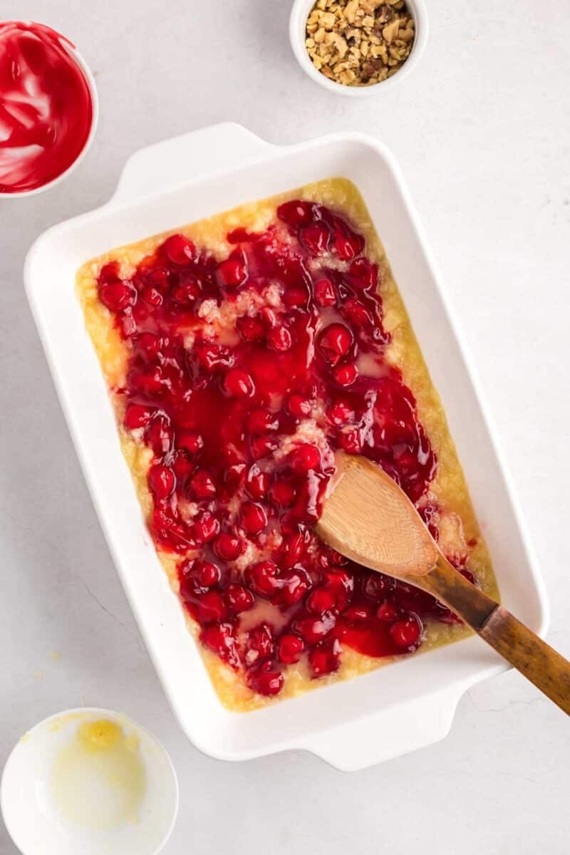 A white baking dish with cherry pie filling and pineapple mixture being stirred with a wooden spoon, surrounded by bowls of chopped nuts and whipped topping.
