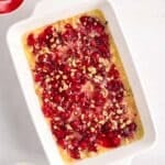 Rectangular baking dish filled with an unbaked cherry dump cake, topped with cherry pie filling and chopped nuts. Mixing bowls with ingredients are nearby on a white surface.
