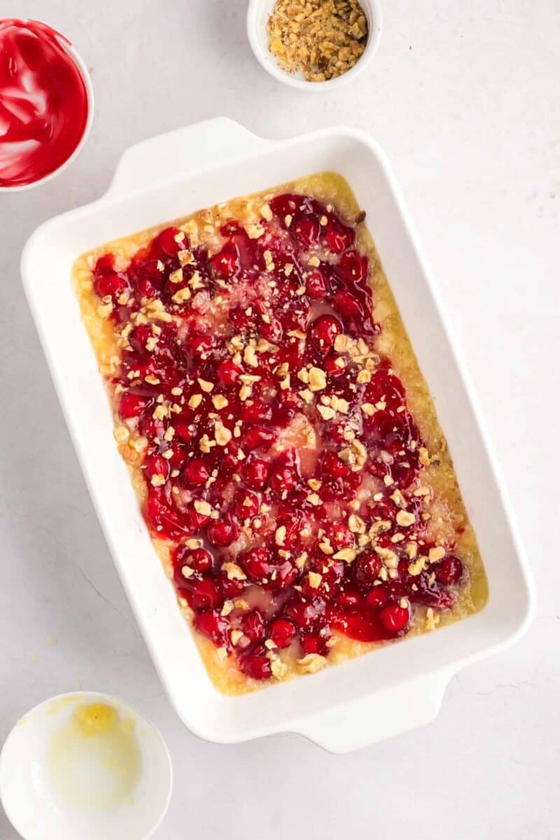 Rectangular baking dish filled with an unbaked cherry dump cake, topped with cherry pie filling and chopped nuts. Mixing bowls with ingredients are nearby on a white surface.