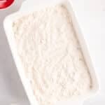 A white baking dish filled with a layer of dry flour mixture sits on a light surface, surrounded by small bowls containing chopped nuts, a red substance, and a yellow liquid.