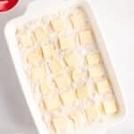 A white baking dish filled with flour mixture topped with evenly spaced butter slices, surrounded by small bowls with ingredients on a white surface.