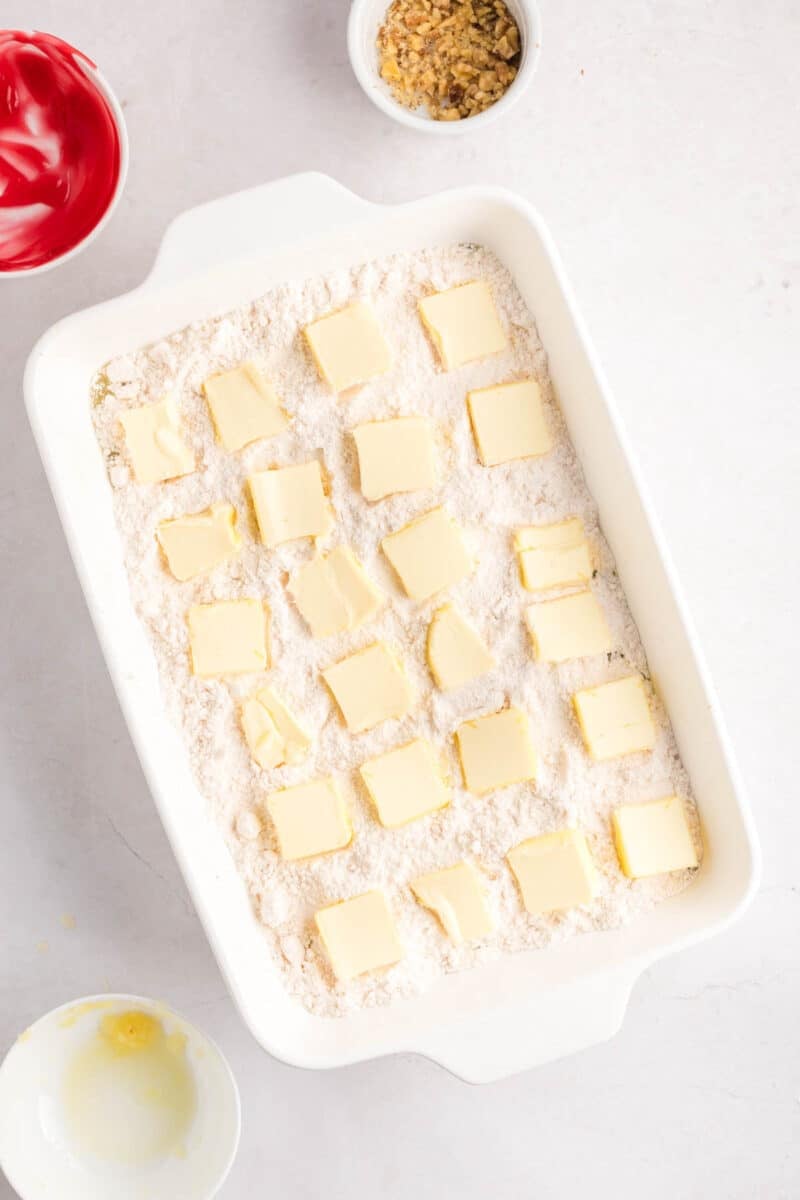 A white baking dish filled with flour mixture topped with evenly spaced butter slices, surrounded by small bowls with ingredients on a white surface.