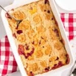 A rectangular baking dish of cherry pineapple dump cake with a golden, crumbly topping and a serving spoon, set on a red checkered cloth. Plates and utensils are nearby.