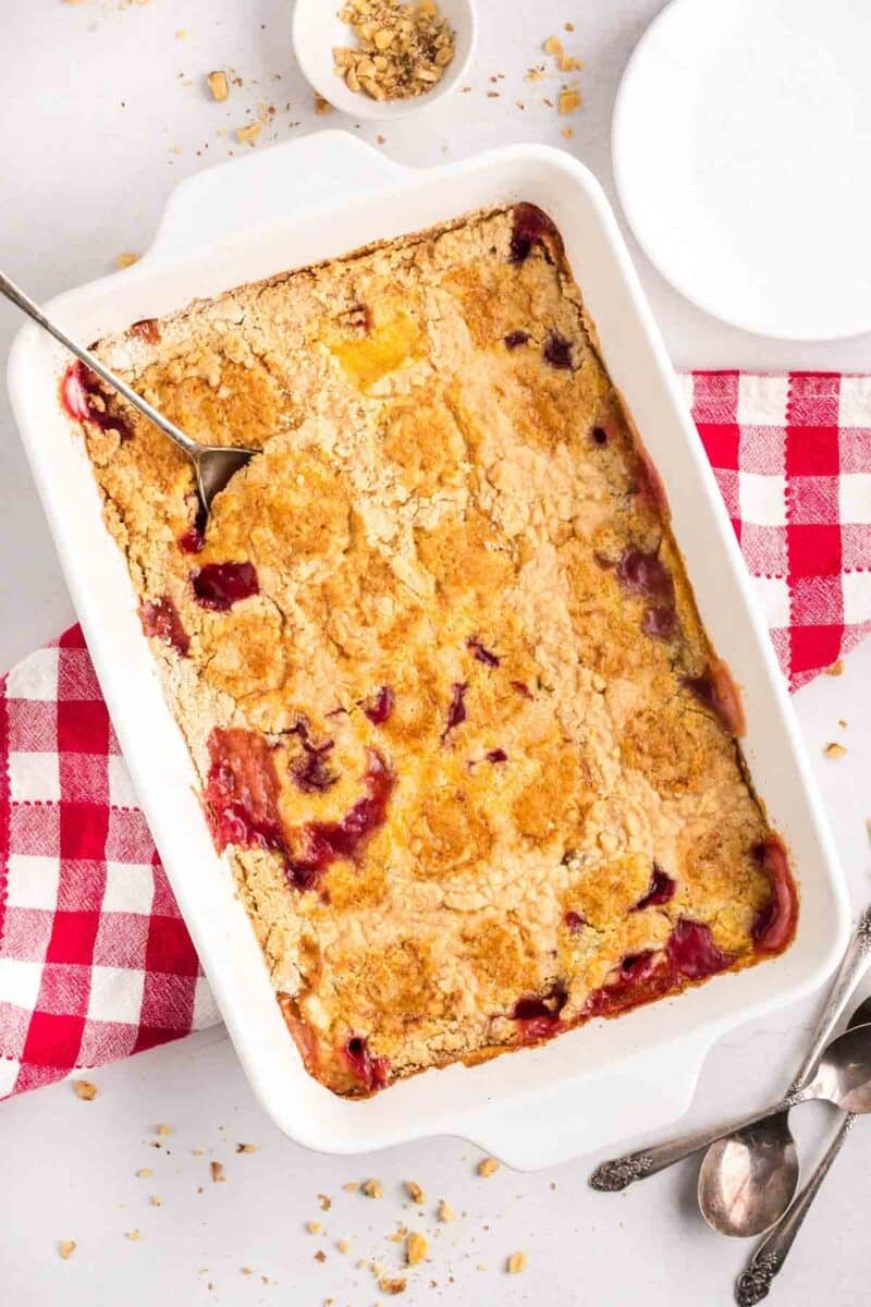 A rectangular baking dish of cherry pineapple dump cake with a golden, crumbly topping and a serving spoon, set on a red checkered cloth. Plates and utensils are nearby.