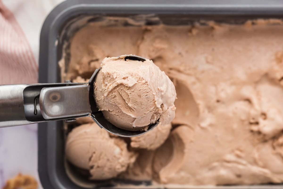 An ice cream scoop holding a ball of chocolate ice cream above a container filled with more chocolate ice cream.