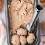 Rectangular metal container filled with chocolate ice cream, with several scoops at one end and an ice cream scoop resting in the container. Waffle cone pieces and a towel are nearby.