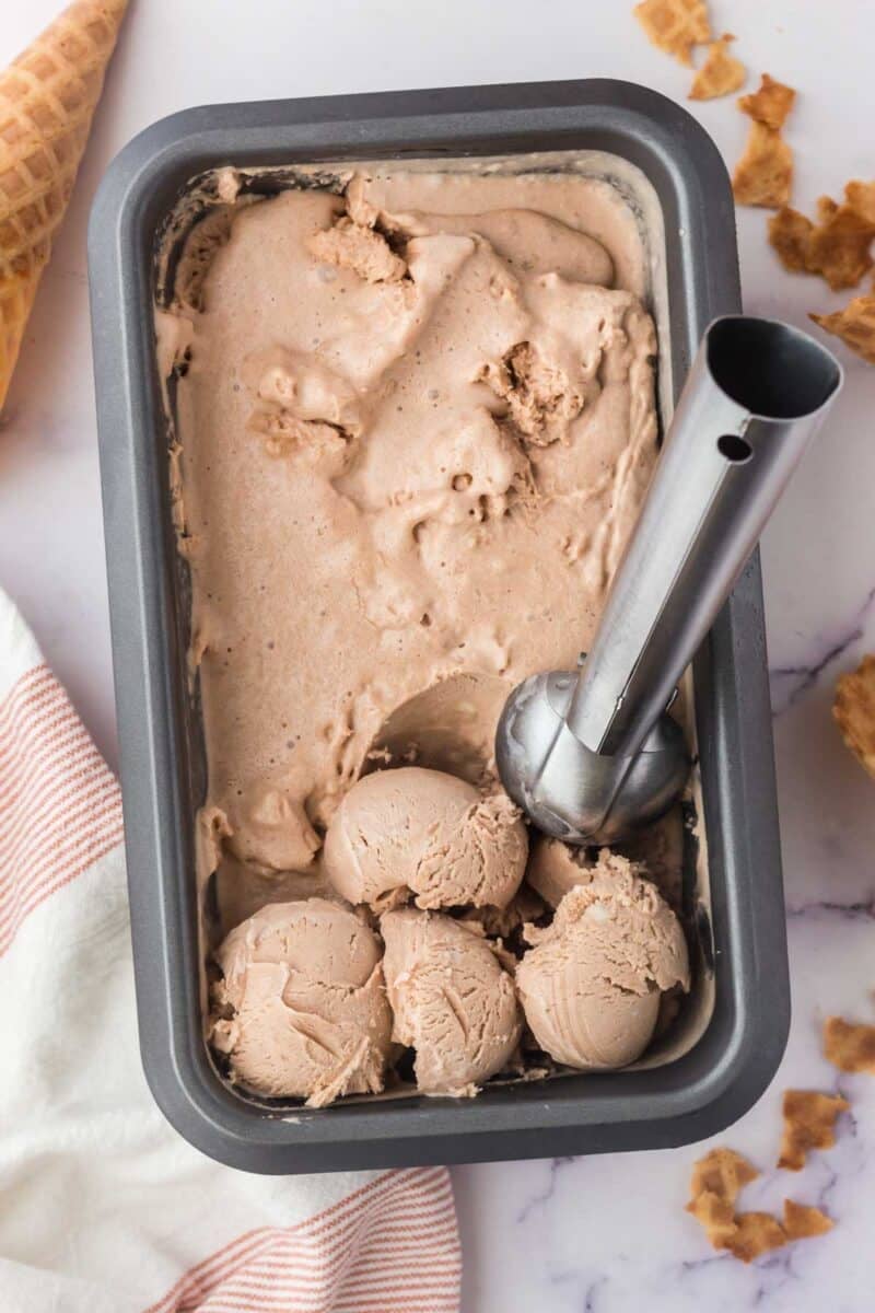 Rectangular metal container filled with chocolate ice cream, with several scoops at one end and an ice cream scoop resting in the container. Waffle cone pieces and a towel are nearby.