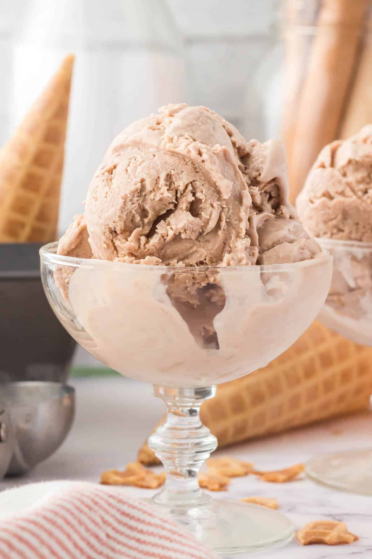 A glass dish filled with several scoops of light brown ice cream, with waffle cones and an ice cream scoop in the background.