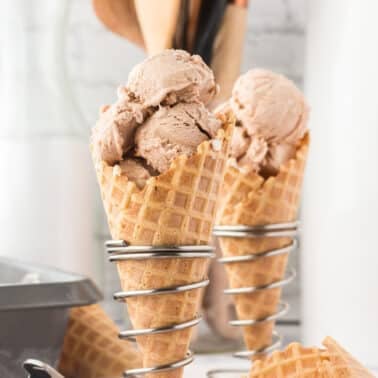 Two waffle cones filled with scoops of light brown ice cream are placed in metal cone holders on a white surface, with kitchen utensils in the background.