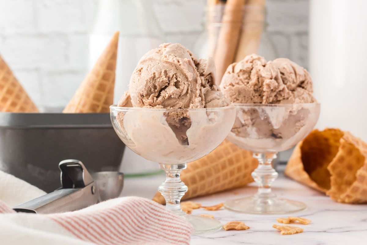 Two glass dessert cups filled with scoops of chocolate ice cream are placed on a counter, surrounded by waffle cones, an ice cream scoop, and crumbs.