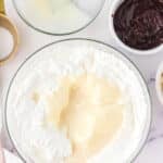 A large bowl of whipped cream mixed with sweetened condensed milk, surrounded by bowls of melted chocolate, more condensed milk, and a measuring cup on a marble surface.