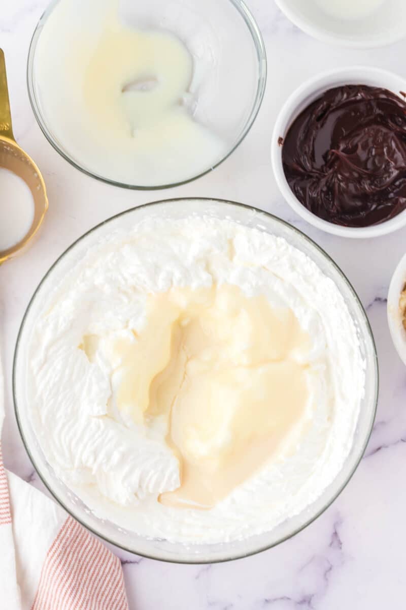 A large bowl of whipped cream mixed with sweetened condensed milk, surrounded by bowls of melted chocolate, more condensed milk, and a measuring cup on a marble surface.