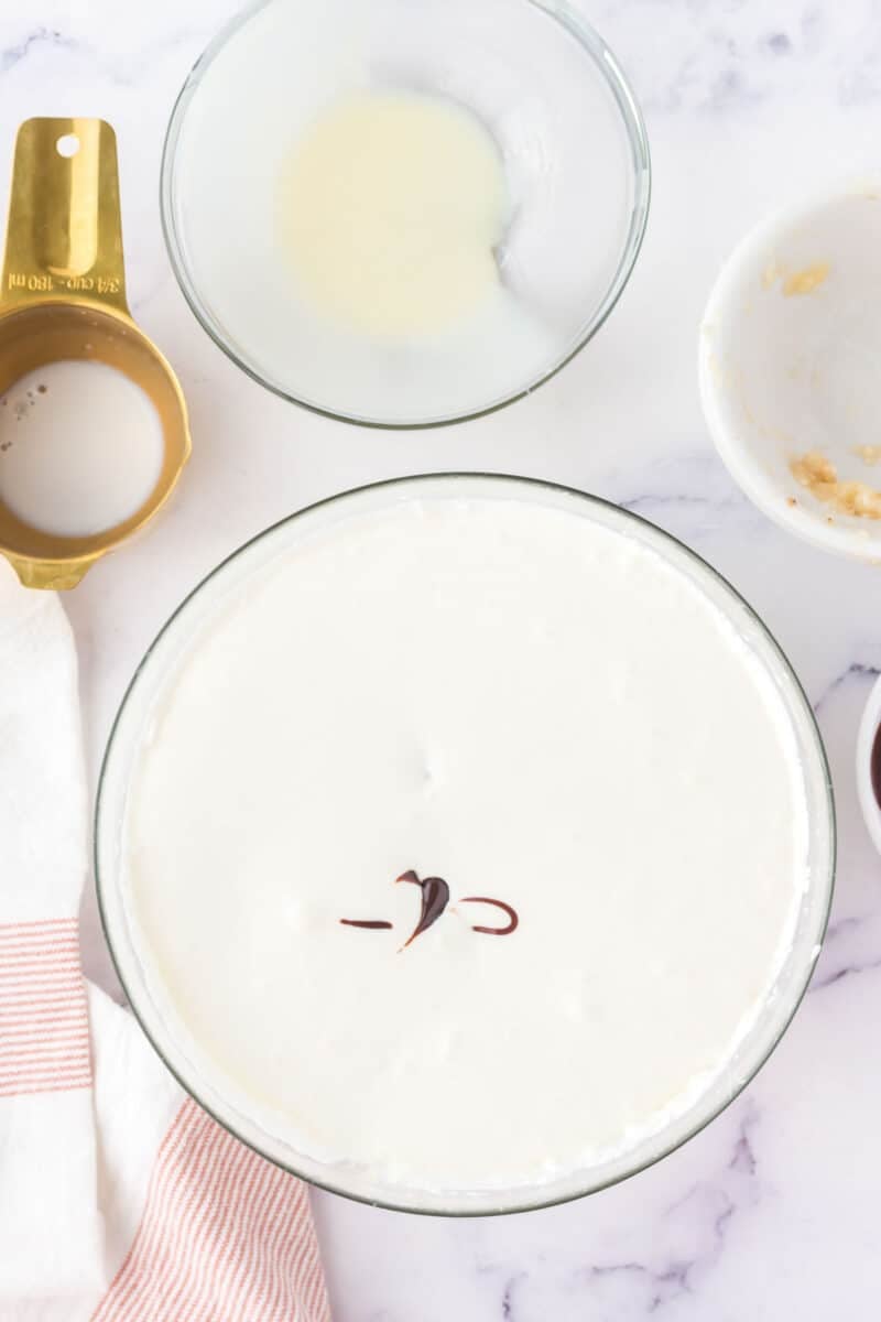 A large bowl of creamy white mixture with a small swirl of chocolate on top, surrounded by empty bowls, a measuring cup, and a striped kitchen towel on a marble surface.